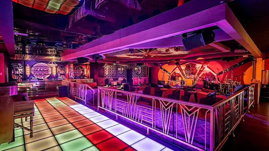 Interior of a nightclub with a lit multicolor dance floor, art deco railings and a stage with a drum kit under purple and orange lighting