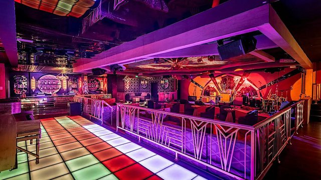 Interior of a nightclub with a lit multicolor dance floor, art deco railings and a stage with a drum kit under purple and orange lighting