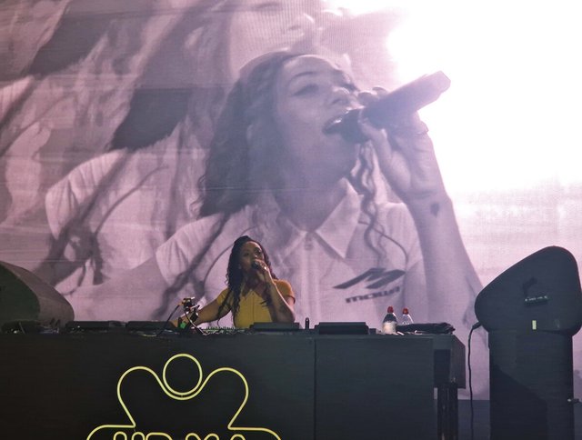 Nia Archives singing into a microphone behind DJ decks, with a large video screen showing her face behind her