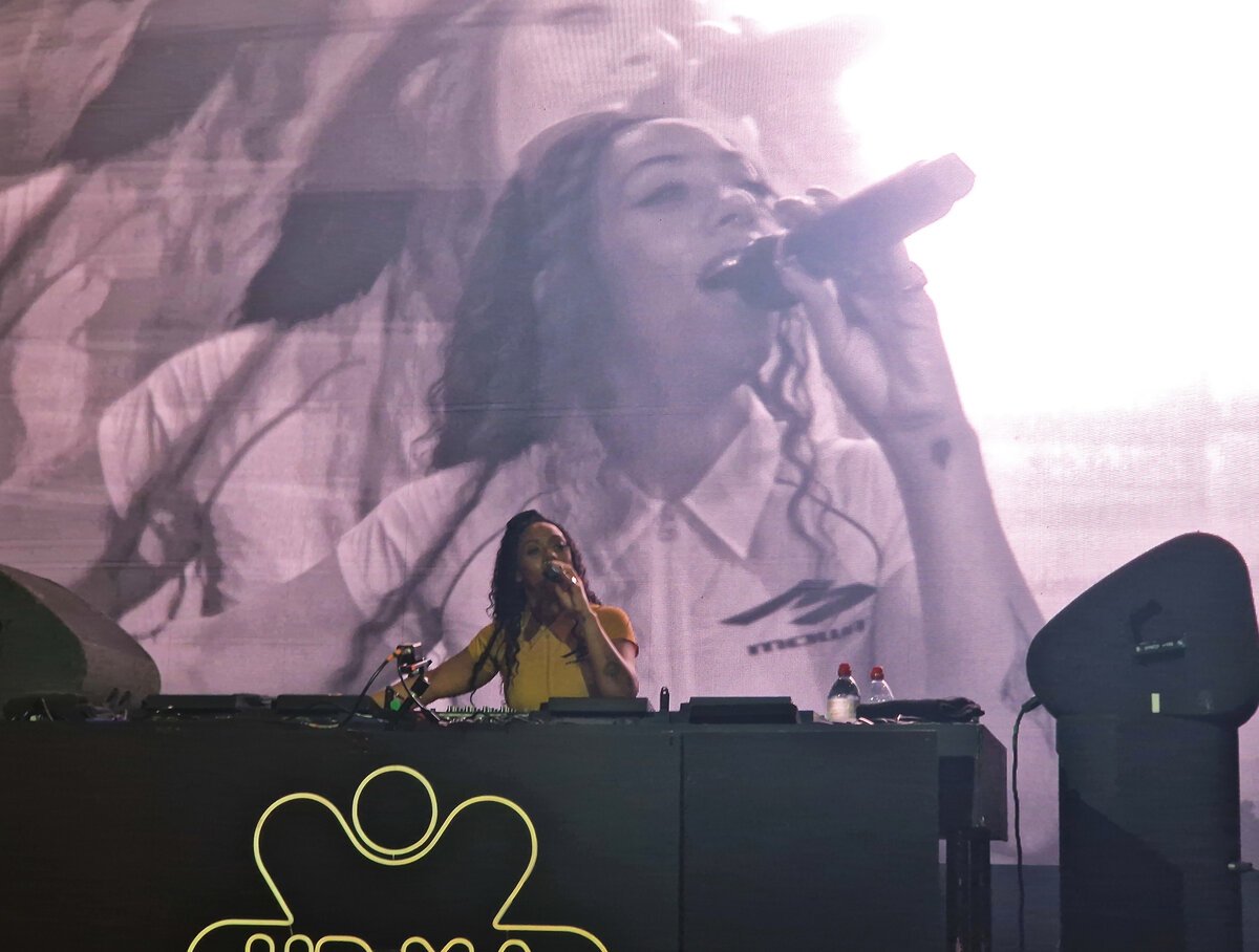 Nia Archives singing into a microphone behind DJ decks, with a large video screen showing her face behind her
