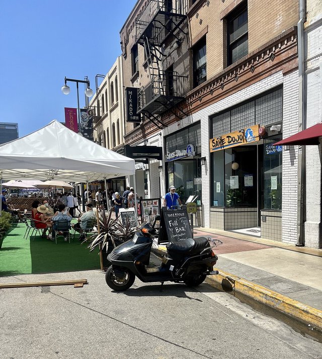 Sunny Little Tokyo street with outdoor seating under a white canopy and a parked scooter in front of a sake bar