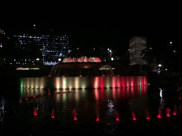 Illuminated fountain glowing red and green at night with lit office buildings in the dark behind it