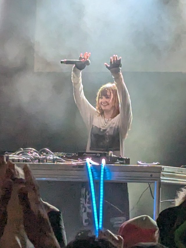 DJ with both arms raised behind a Pioneer DJ setup in stage haze, with a blue light strip in front of the booth