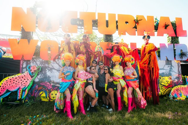 Costumed Nocturnal Wonderland performers in neon wigs and stilts posing with festivalgoers by the painted festival sign