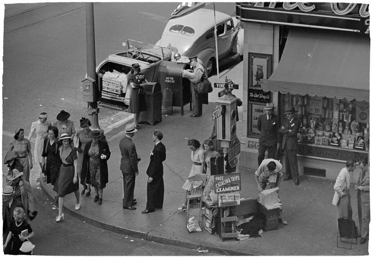 Black-and-white 1940 street corner at Hollywood and Vine, with pedestrians, a mail carrier, a parked convertible and a drugstore awning