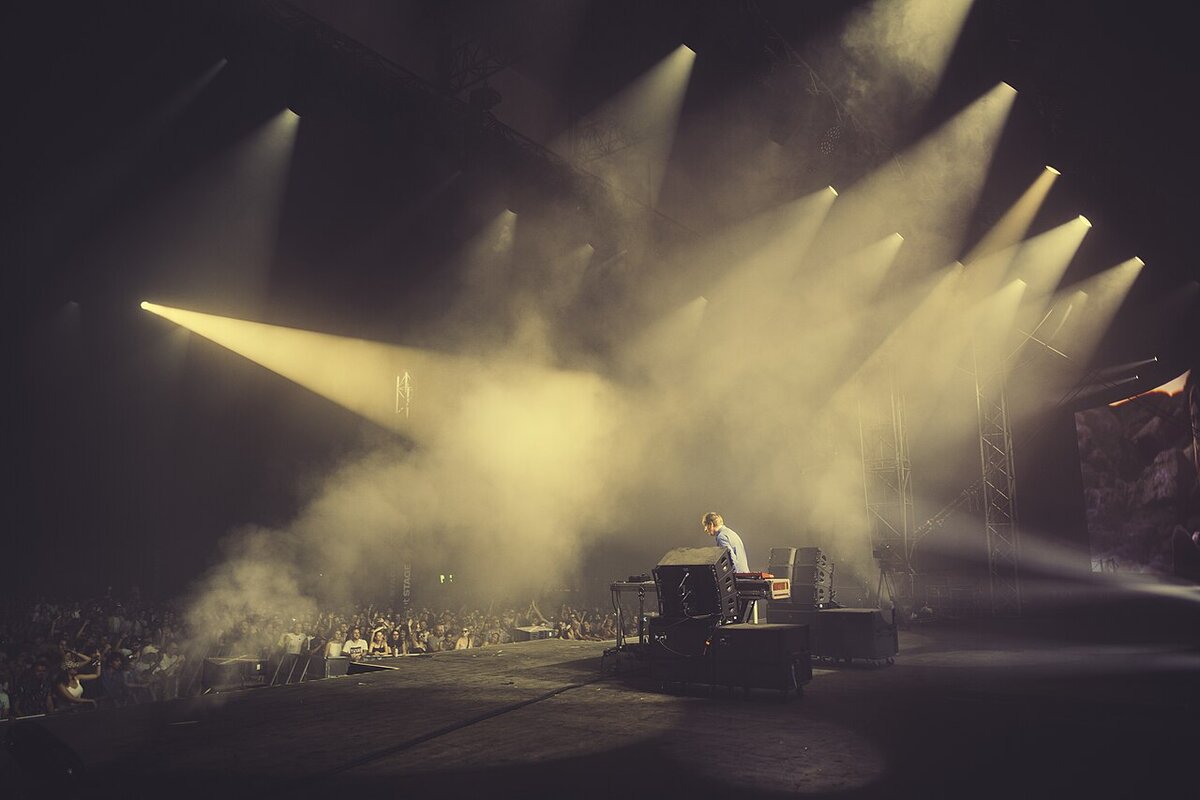 Lone DJ at a stage setup facing a crowd, with shafts of yellow light cutting through haze