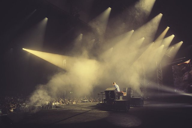 Lone DJ at a stage setup facing a crowd, with shafts of yellow light cutting through haze