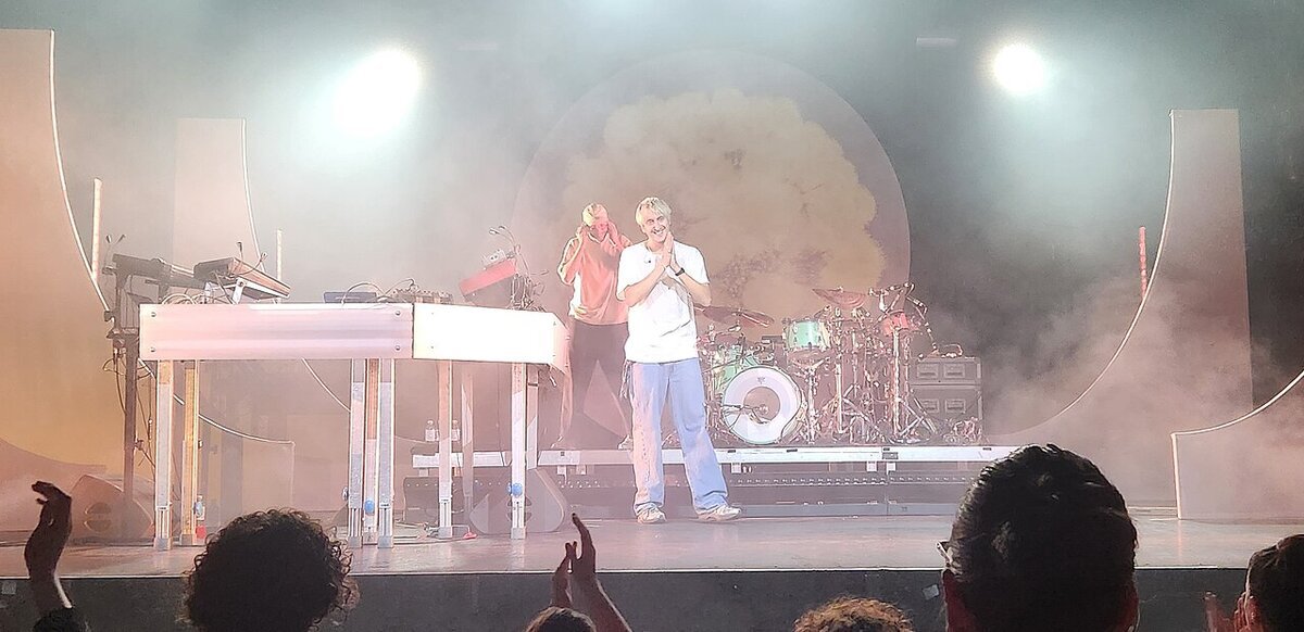 Two performers on a hazy stage with a keyboard rig and drum kit under bright spotlights, with audience heads in the foreground