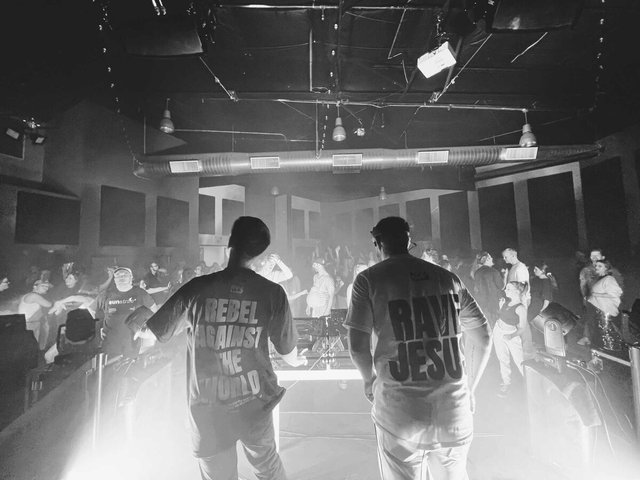 Rave Jesus and a partner behind the decks at a packed club show, seen from behind in matching Rave Jesus shirts