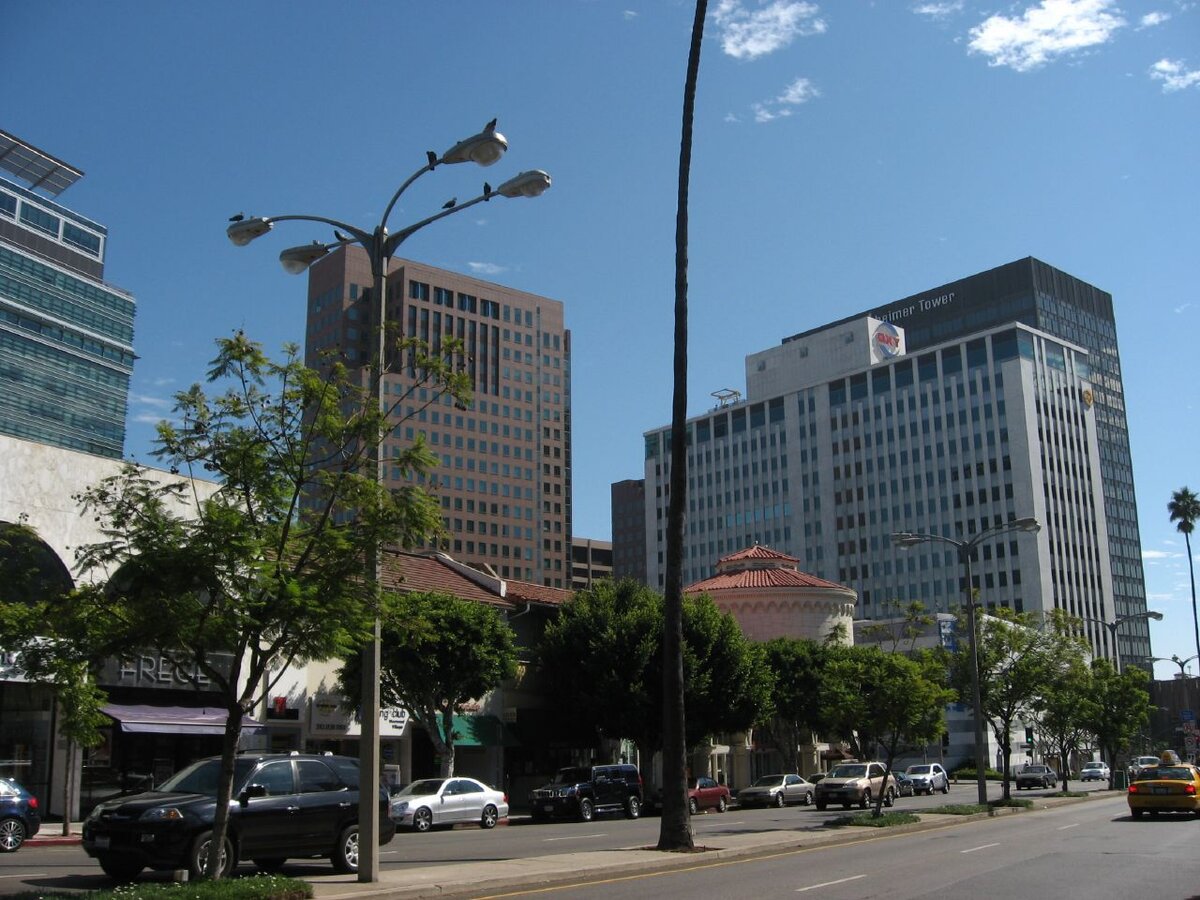 Street view of Westwood Village with office towers, palm trees and passing cars under a blue sky
