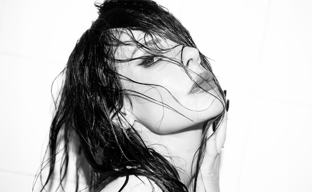 Black and white portrait of a dark-haired woman with wet hair across her face, shown in profile against a white tiled wall