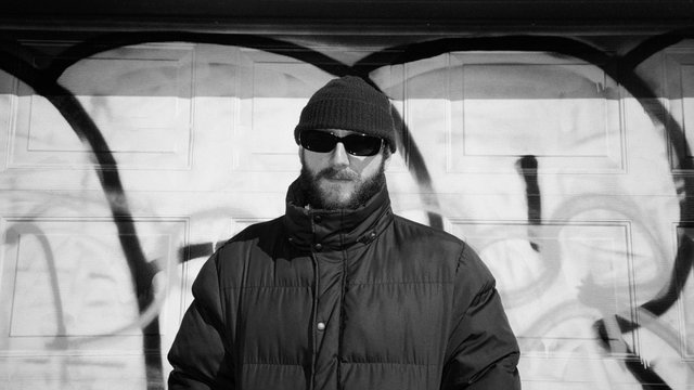 Black and white photo of River Tiber in a beanie, dark sunglasses and a puffer jacket, standing in front of a graffiti-tagged garage door