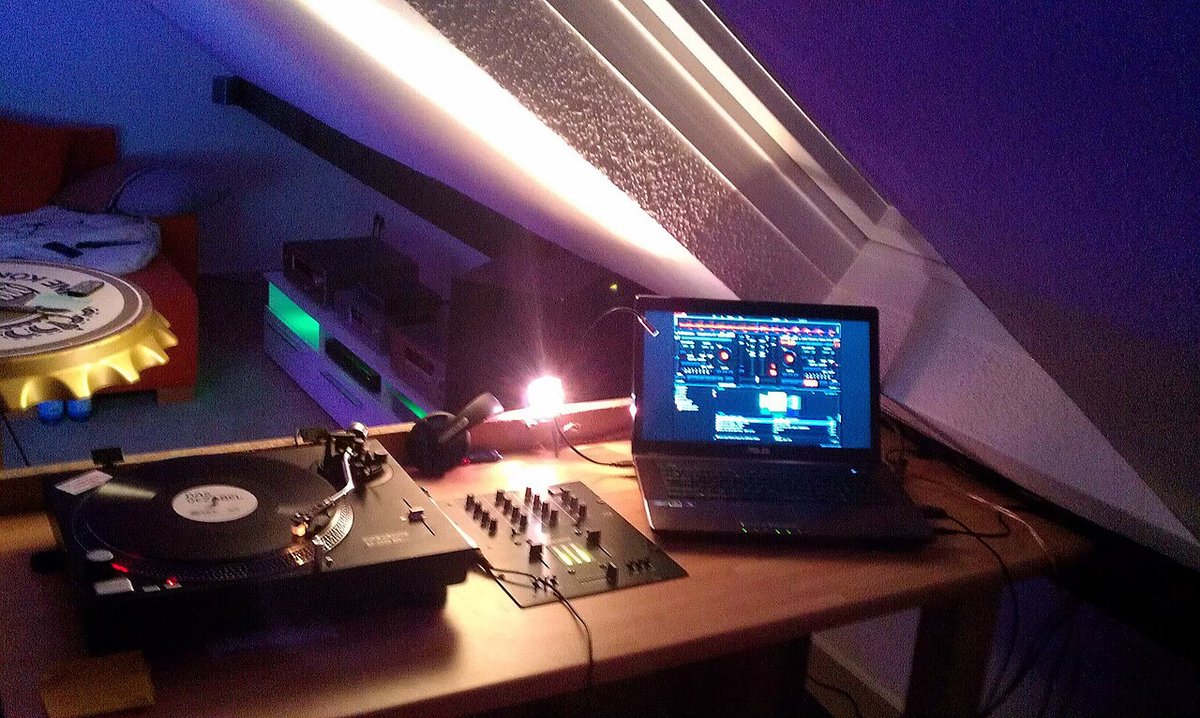 A compact DJ setup with a turntable, mixer and laptop under a slanted ceiling lit by a bright strip light