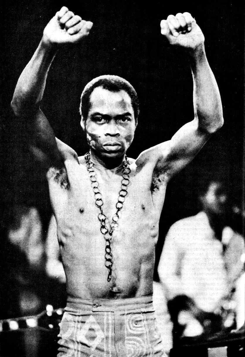Black and white photo of Fela Kuti, shirtless with a chain necklace, raising both clenched fists above his head