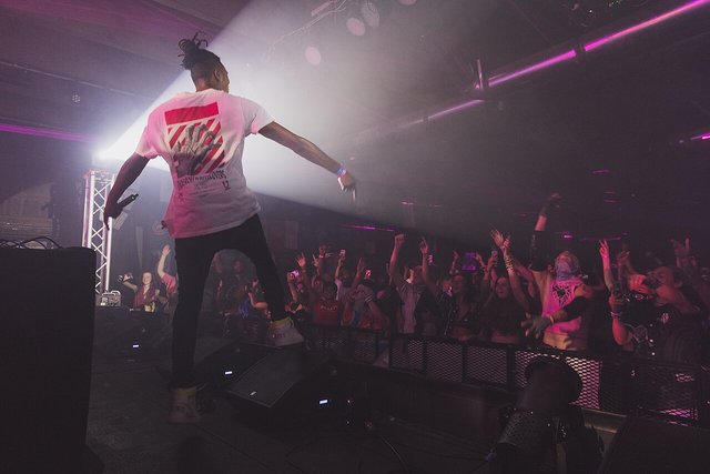 SAYMYNAME standing on stage with arms spread in front of a crowd with raised hands under pink and white club lighting