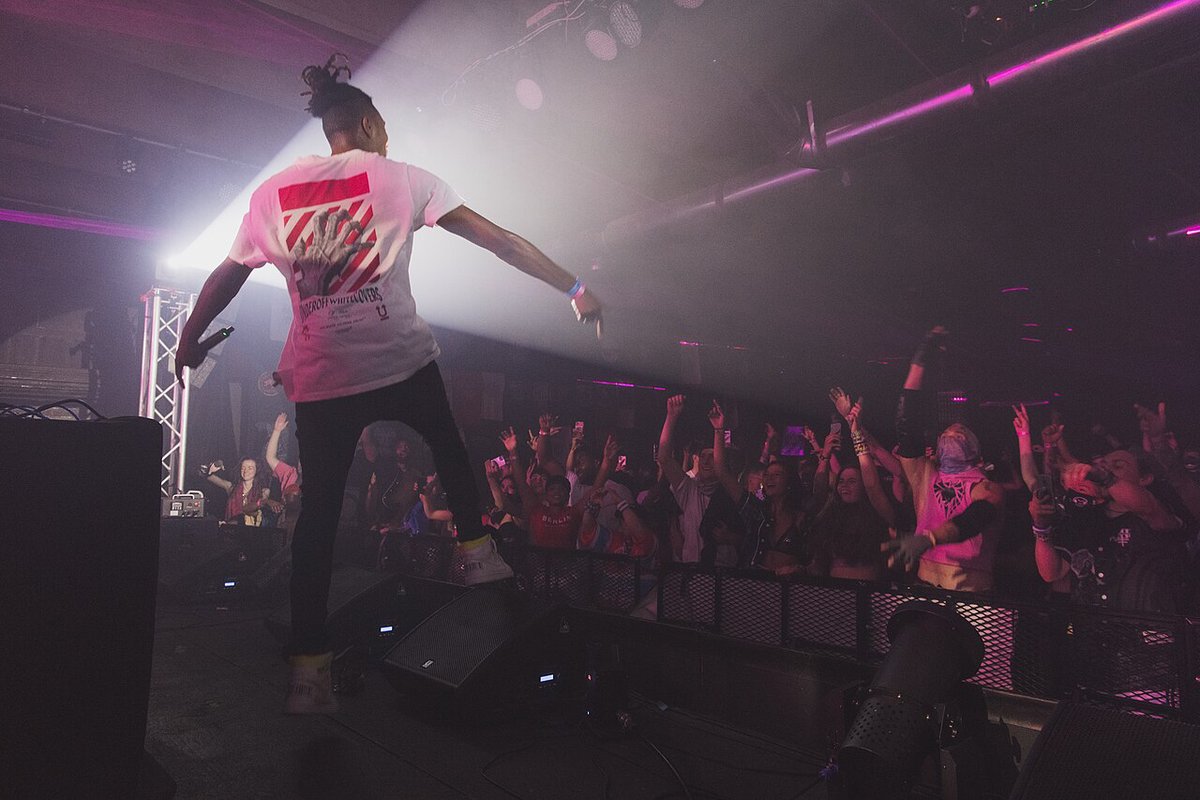 SAYMYNAME standing on stage with arms spread in front of a crowd with raised hands under pink and white club lighting