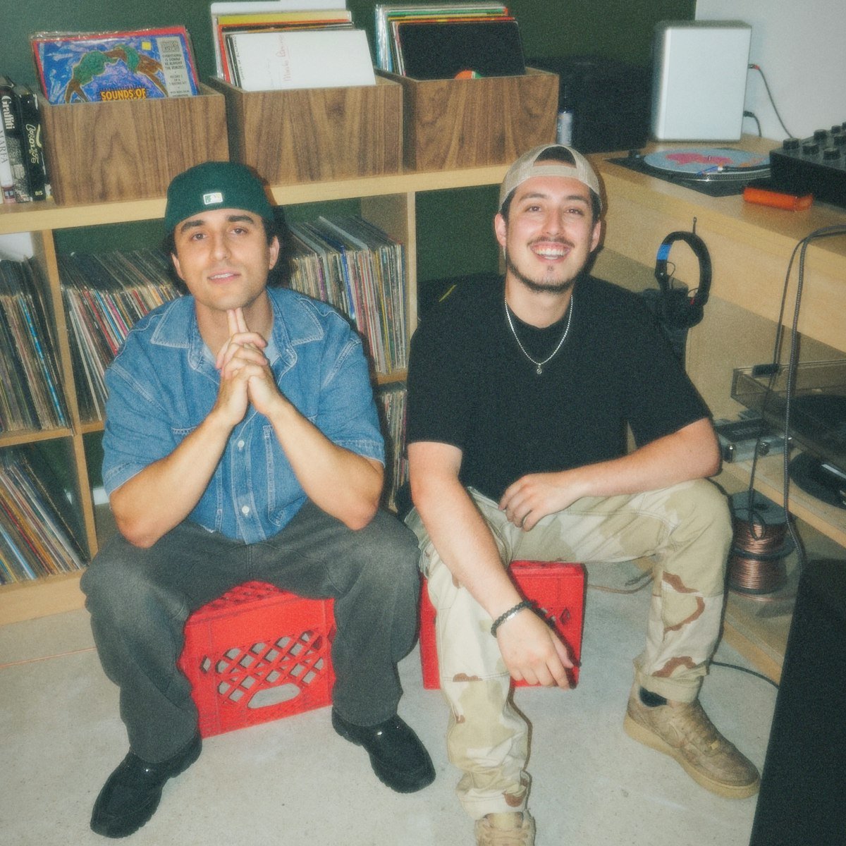 Two members of Slamm seated on red milk crates in a room lined with vinyl records