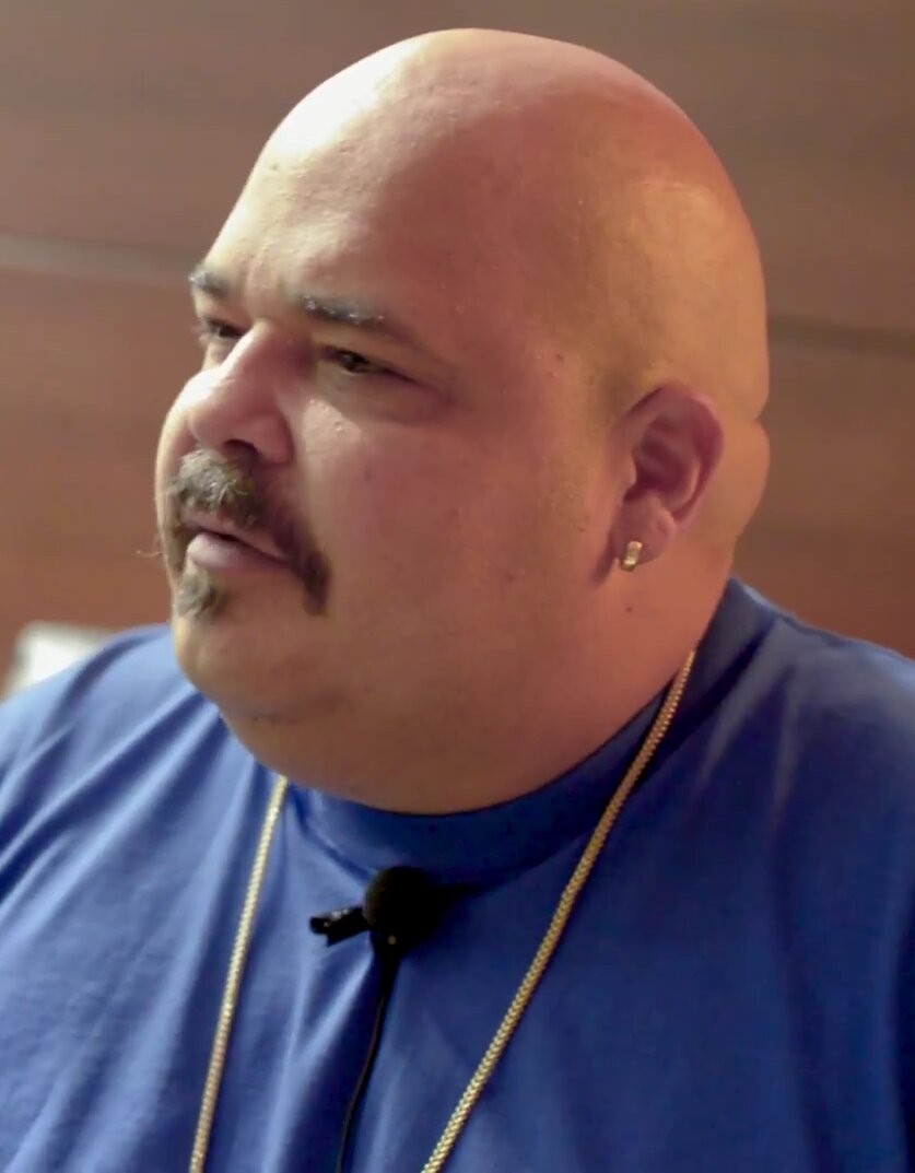 Close-up portrait of DJ Sneak in a blue shirt with a clip-on microphone