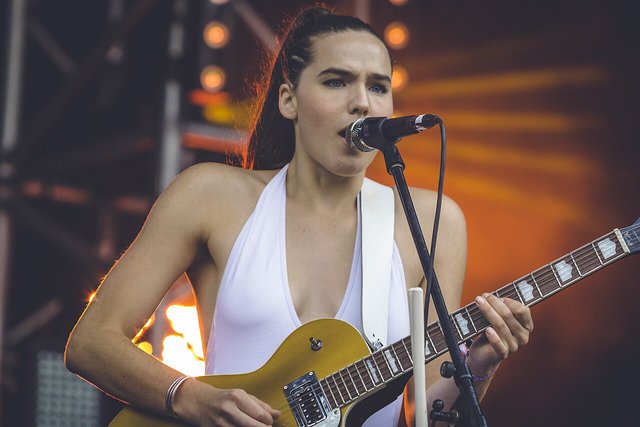 Sophie Hawley-Weld of Sofi Tukker singing into a microphone and playing a gold electric guitar under orange stage light