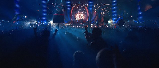 Crowd with hands raised facing a lit electronic music stage with a burst of red light and blue light columns