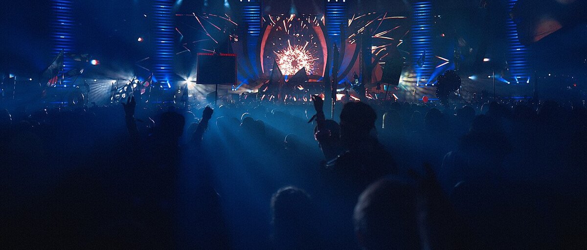 Crowd with hands raised facing a lit electronic music stage with a burst of red light and blue light columns