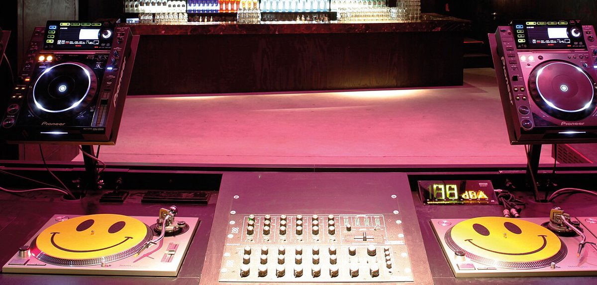A DJ booth with two CDJ players and a mixer, each deck topped with a smiley-face mat