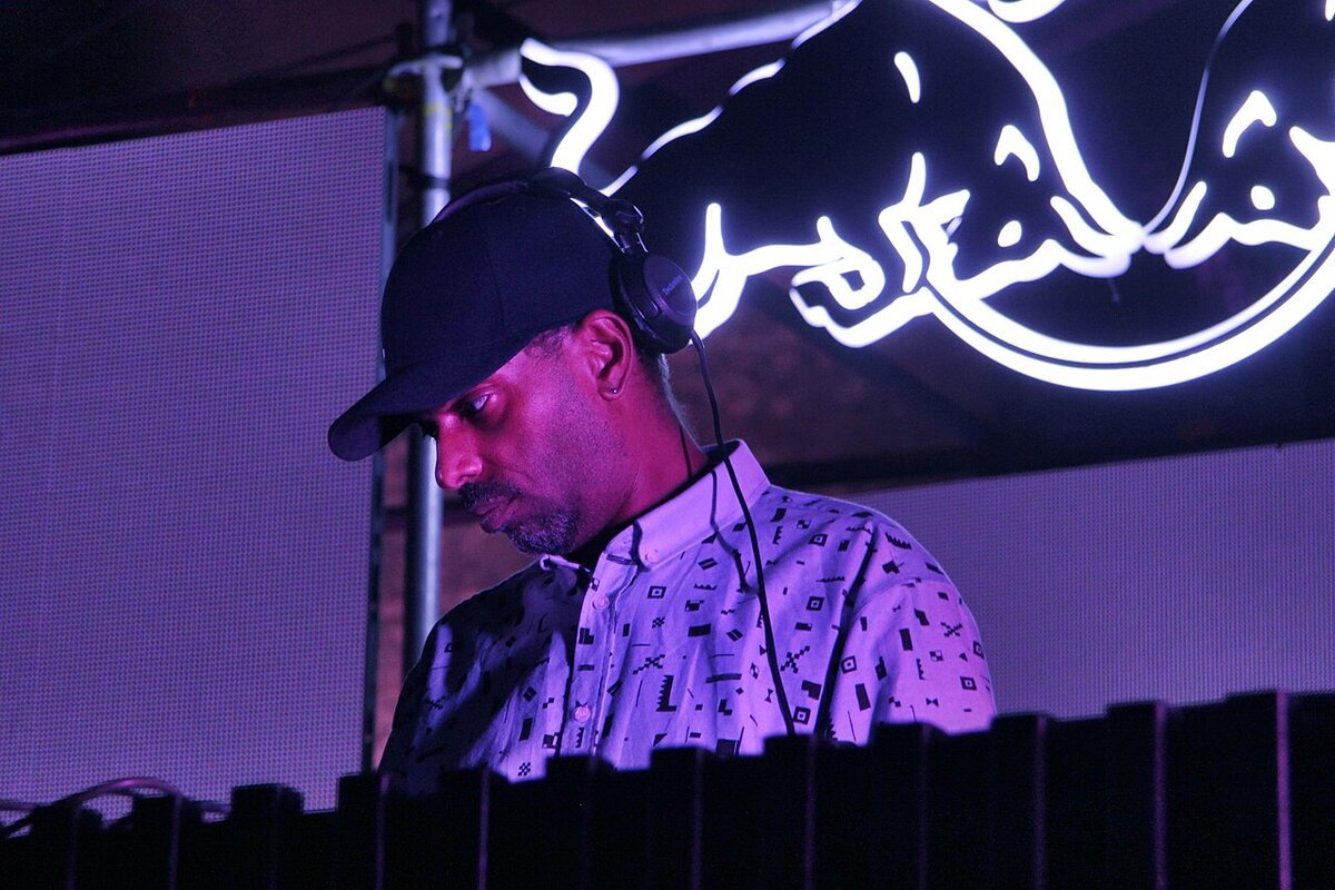 DJ in a dark cap and headphones leaning over the decks under purple light, with a glowing logo sign behind him