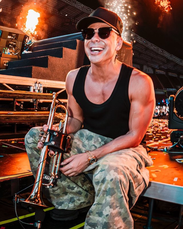 Timmy Trumpet smiling while seated on a stage platform holding his trumpet, with flames and confetti behind him