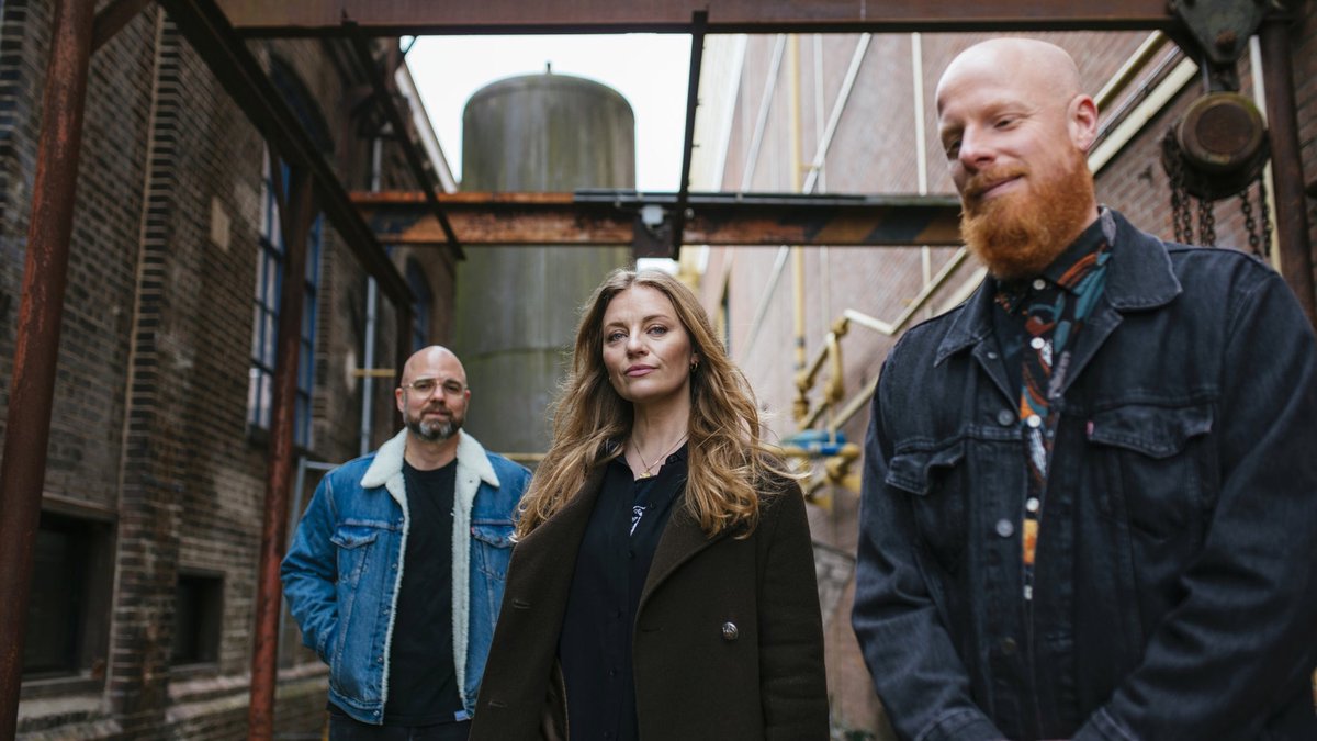 Tinlicker tour photo: three musicians in denim and dark coats standing in a brick industrial courtyard under rusted steel beams