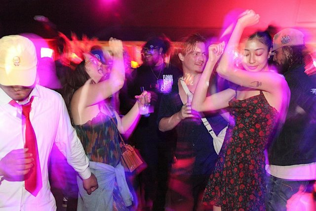 Motion-blurred dancers under pink and purple club lighting, mid-move with drinks in hand