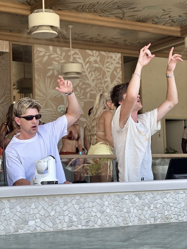 Two DJs with their arms raised behind a tiled booth at a daytime pool party