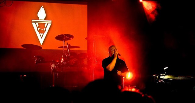 VNV Nation's singer on stage in red stage light beside a screen showing the band's torch logo