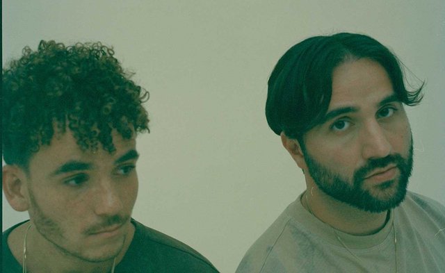 Press photo of two men, one with curly hair and one bearded, against a pale green wall