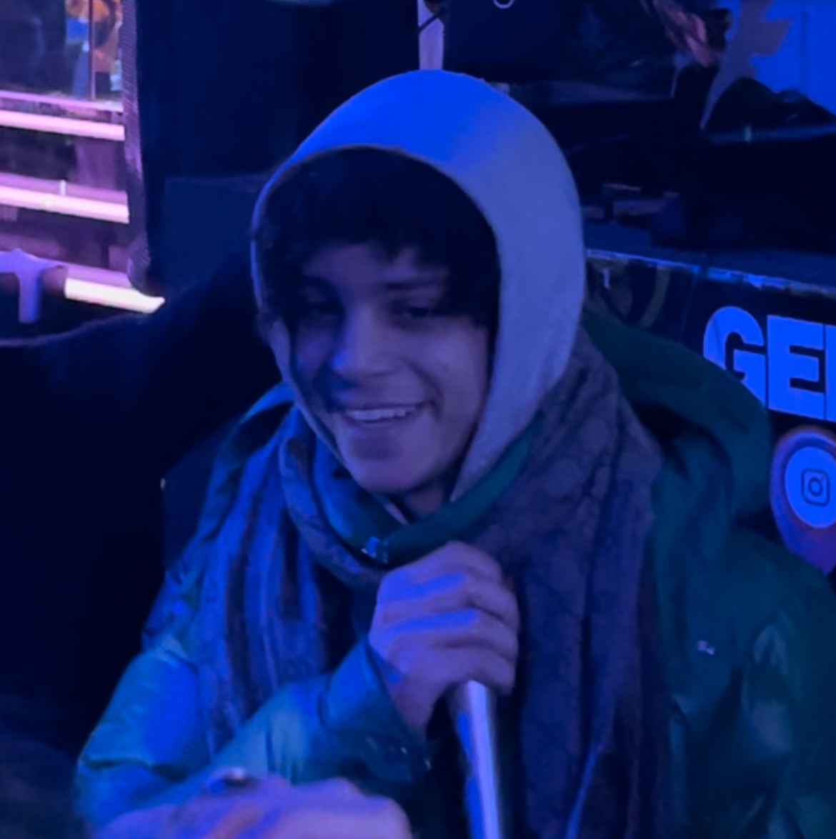 Xaviersobased in a gray hoodie and scarf, smiling and holding a microphone under blue stage light