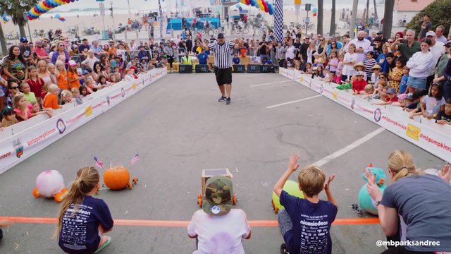 Kids cheering at the starting line of the Manhattan Beach Pumpkin Race as a referee signals and a crowd lines the course