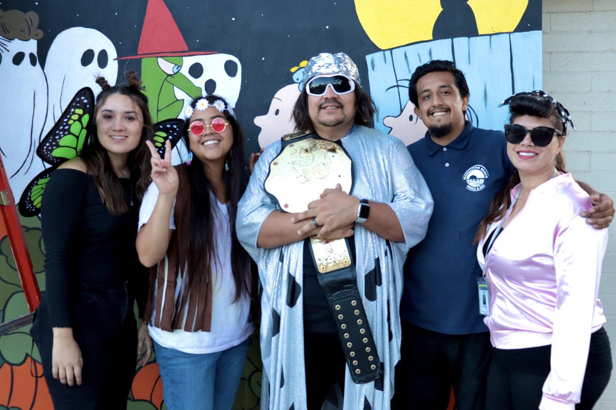 Five costumed staff and volunteers posing in front of a painted Halloween mural with ghosts, a witch hat and a harvest moon