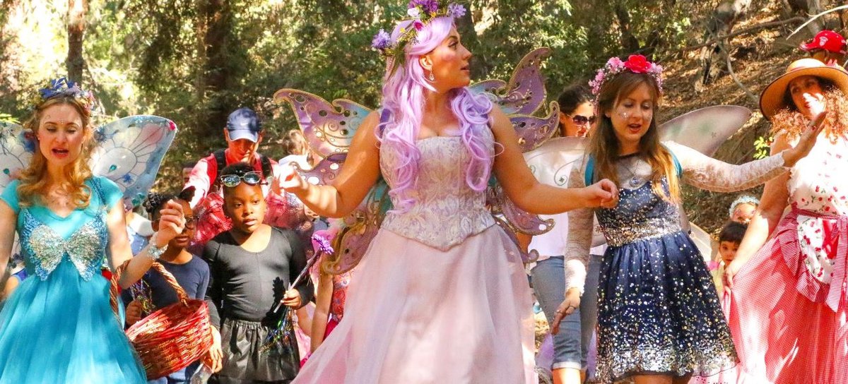 Costumed faeries with wings and flower crowns walking a wooded trail with children carrying baskets
