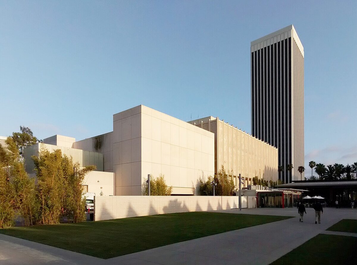 Cream-colored LACMA museum buildings and a tall dark tower beside a lawn at dusk