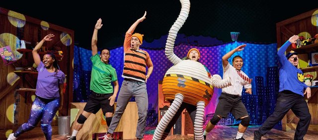 Six cast members in bright costumes strike matching dance poses on a colorful comic-book city stage set