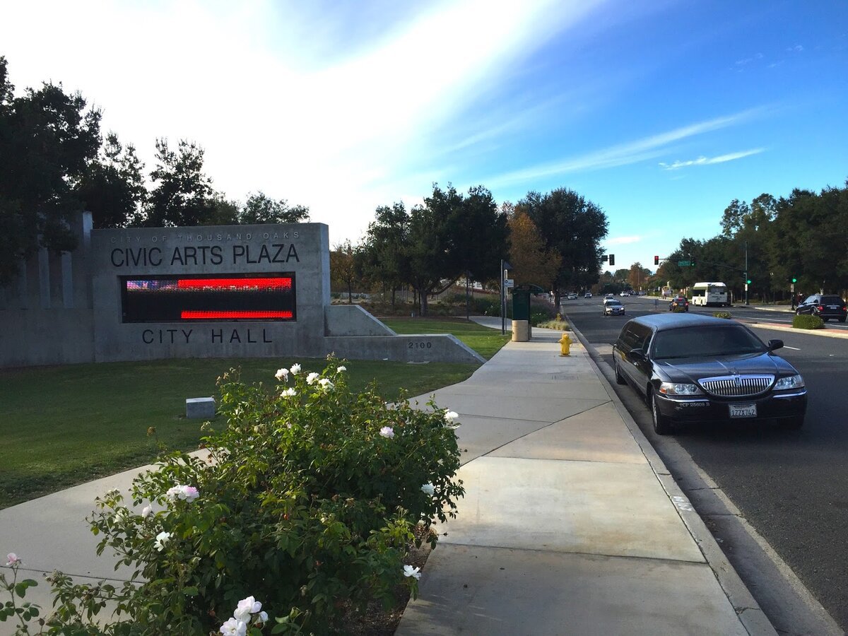 Thousand Oaks Civic Arts Plaza entrance sign beside a sidewalk and tree-lined street under a blue sky