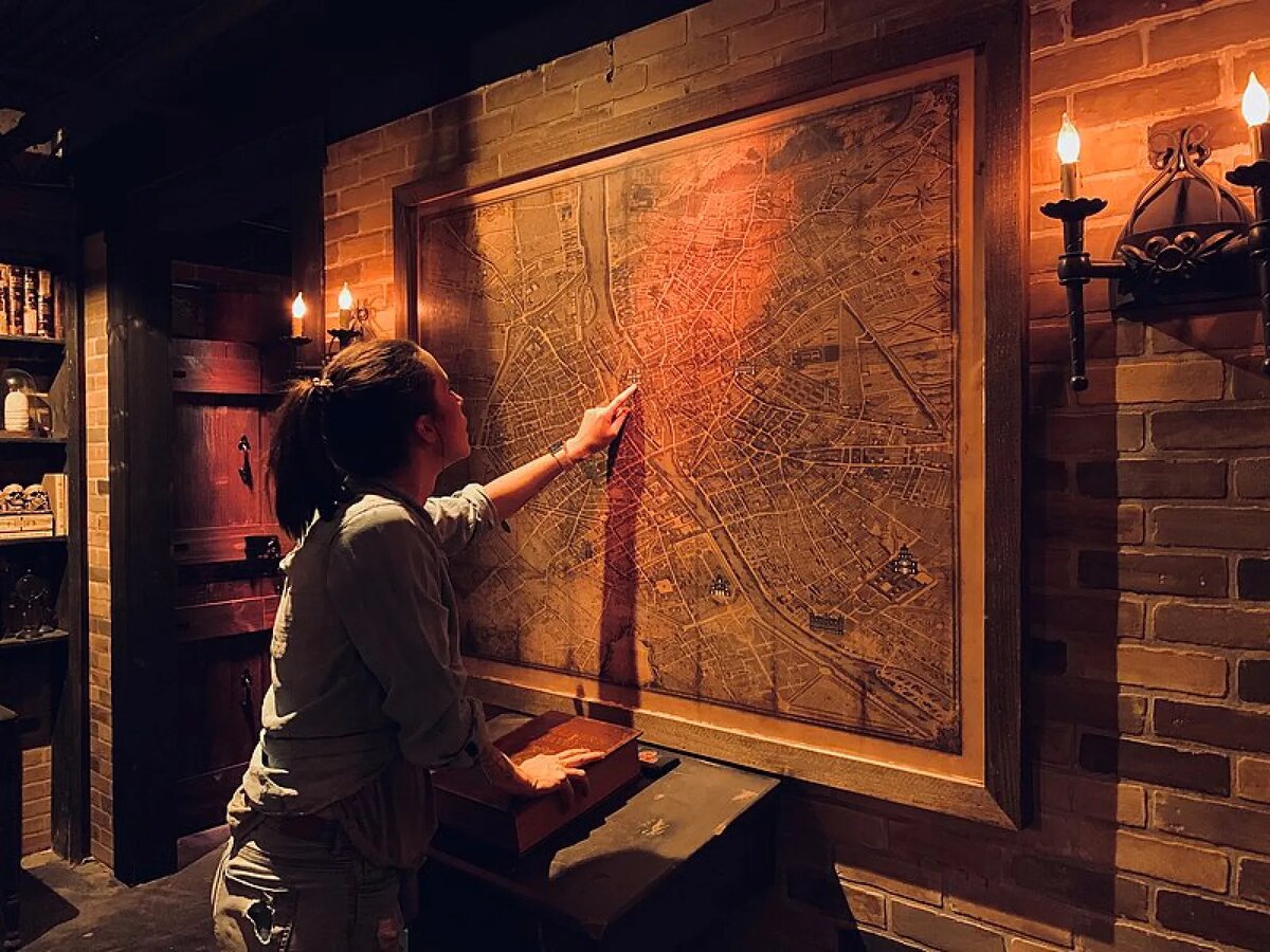 A woman in a dim, candlelit brick room points at a large antique city map in a wooden frame