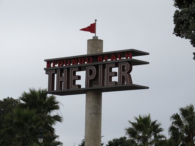 The Redondo Beach The Pier sign on a tall pillar with a red pennant, framed by palm trees under a gray sky