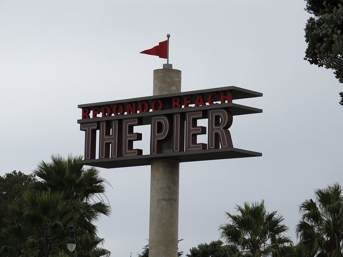 The Redondo Beach The Pier sign on a tall pillar with a red pennant, framed by palm trees under a gray sky