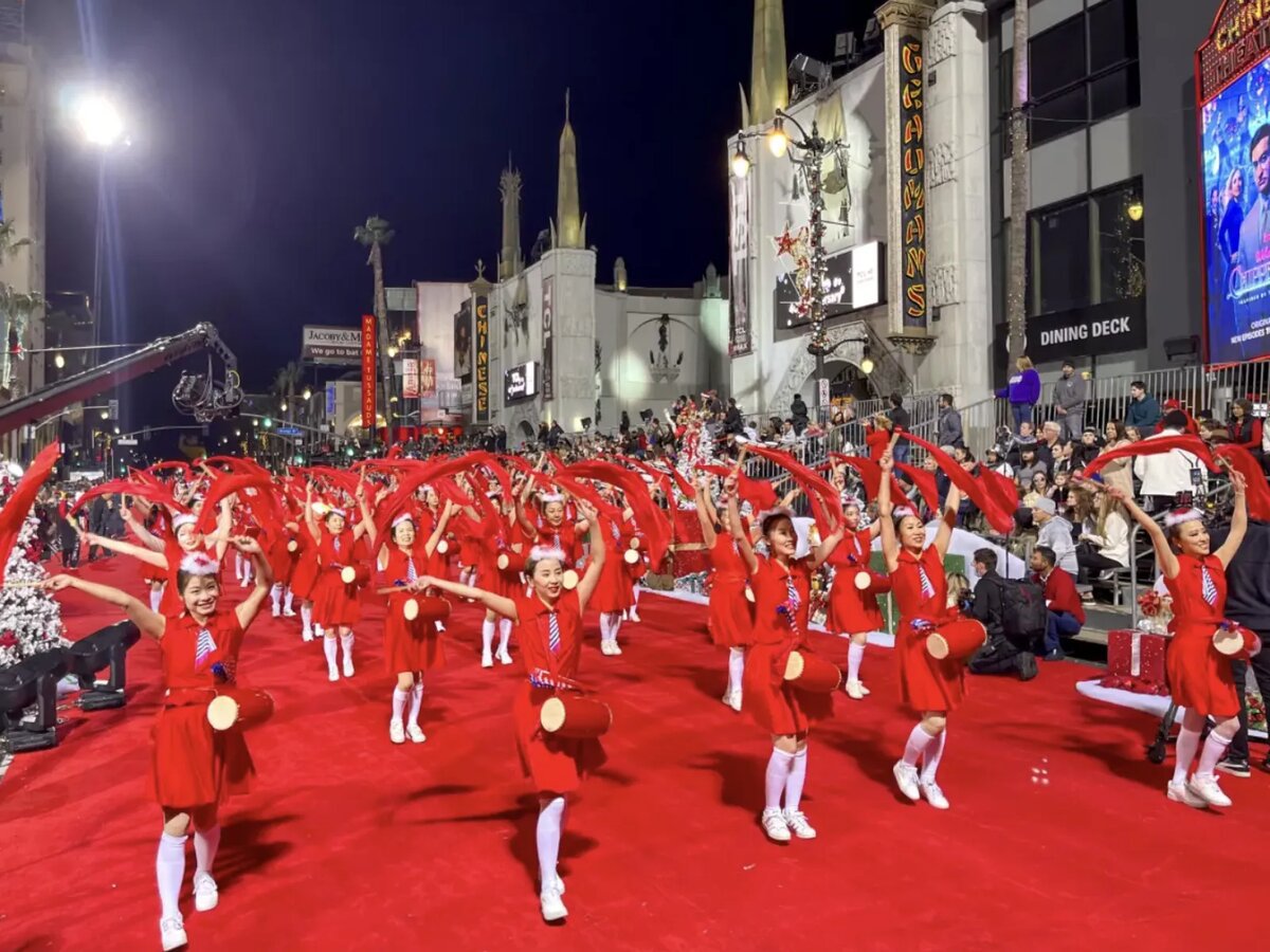 Dancers in red costumes waving ribbons on a red carpet along Hollywood Boulevard during the parade, with the Chinese Theatre behind them