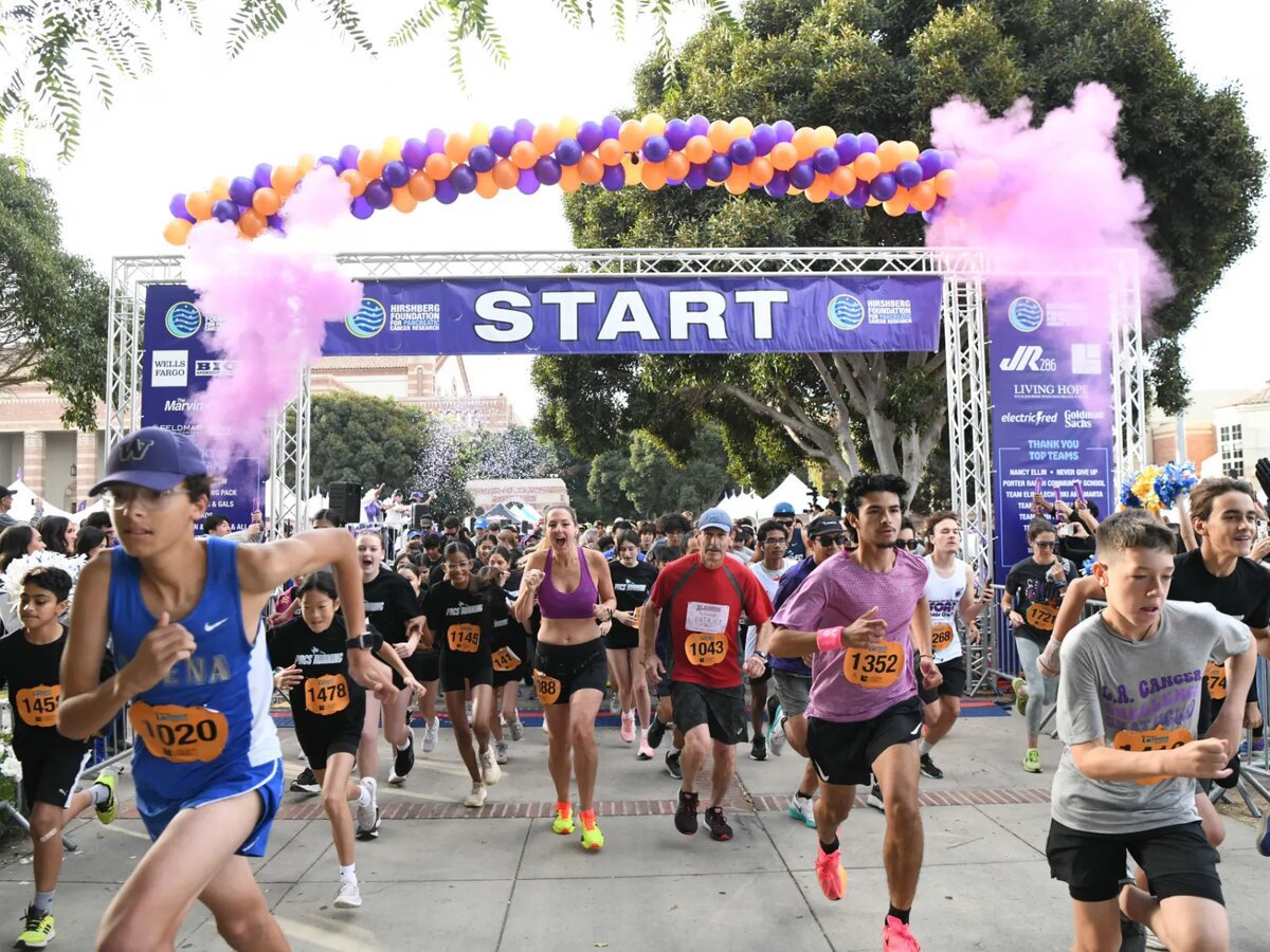 Runners and walkers, some in costume, surge through the purple START arch under an orange and purple balloon garland
