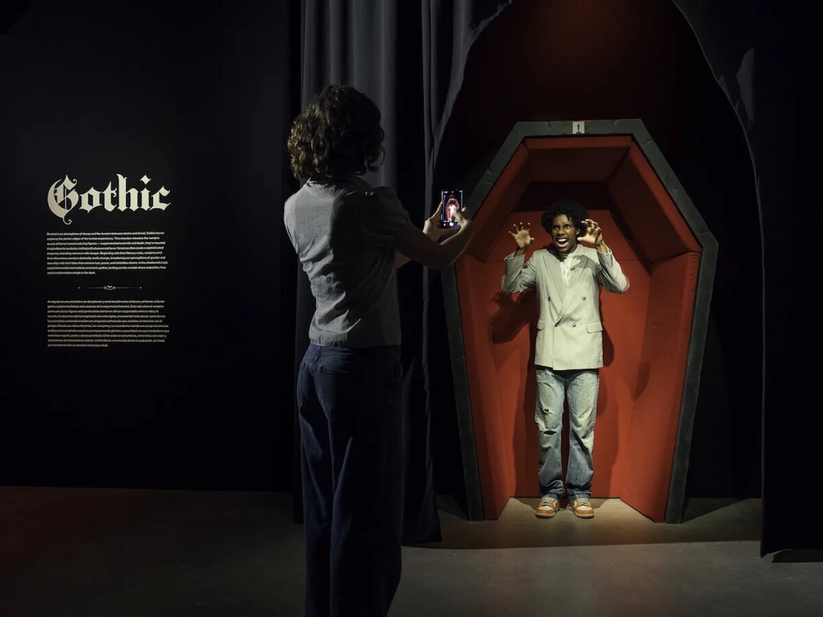 A visitor photographs a person posing with clawed hands inside a red-lined coffin display in a dark gothic gallery