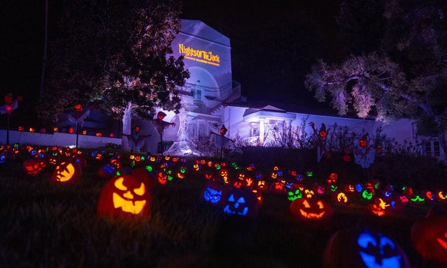 Rows of glowing carved jack-o'-lanterns on a dark lawn in front of the lit white mansion at King Gillette Ranch, with the Nights of the Jack logo projected on its tower
