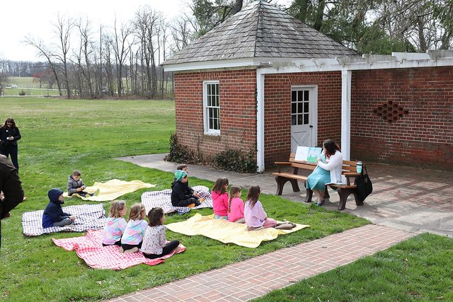A storyteller on a bench holds up an open picture book while a small group of young kids sits on picnic blankets on the grass listening