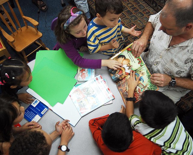 Seen from above, a group of young kids crowds around a library table as an adult holds open a big illustrated picture book