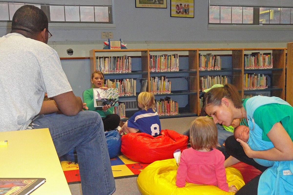 A librarian holds up an open picture book for toddlers sitting on red and yellow beanbags in a children's library corner while parents listen nearby